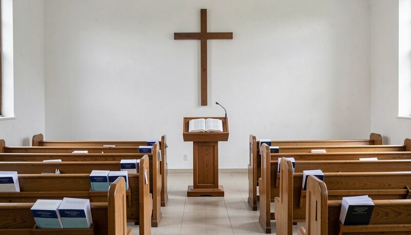 A plain church interior with a wooden cross on a white wall, an open Bible on a lectern and folded worship folders on empty pews.